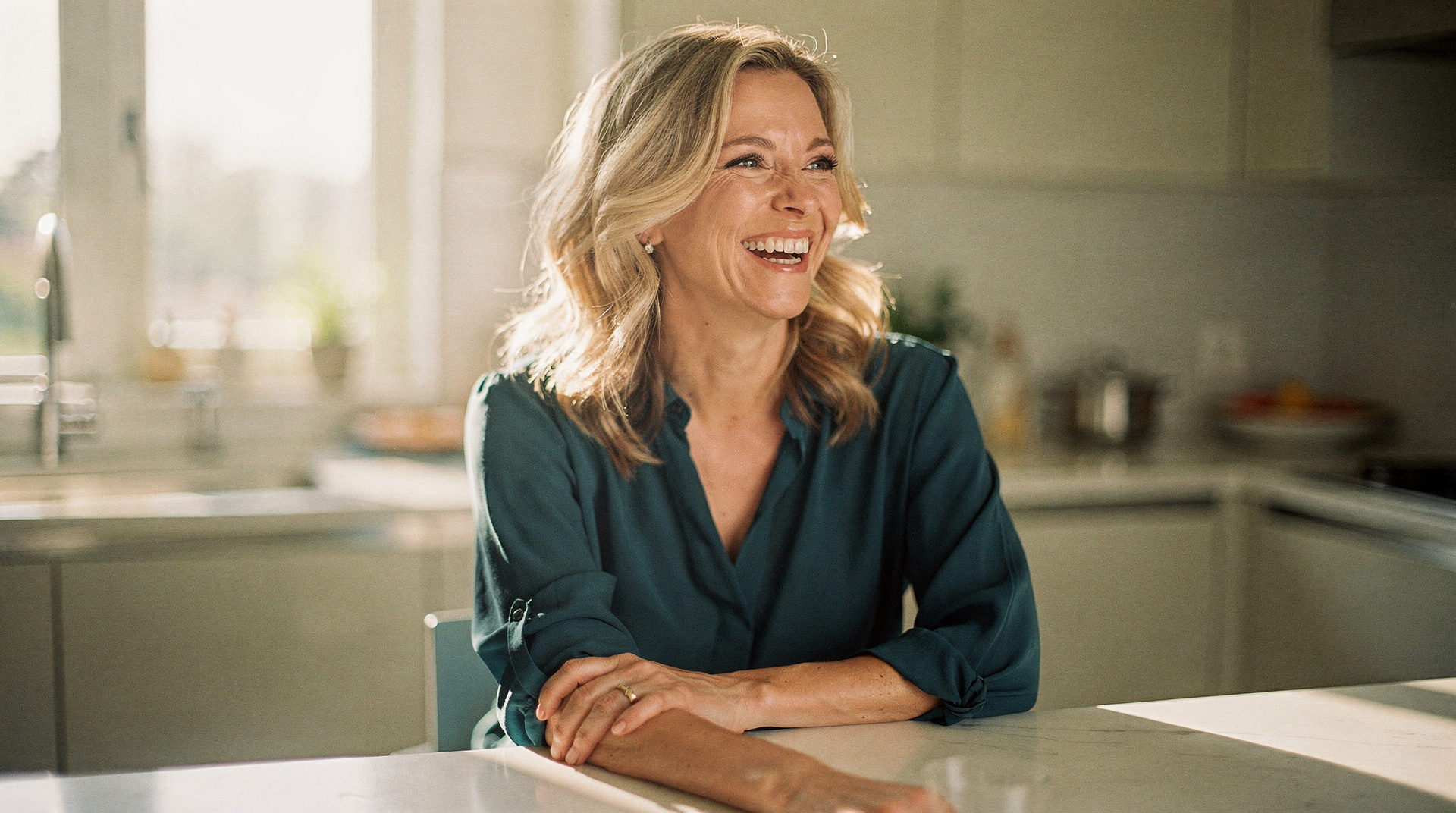 A woman in her mid-forties laughs warmly while leaning on her kitchen counter in morning light