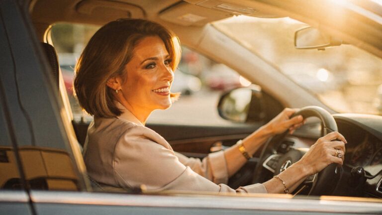 A woman in her late forties sitting in a parked car in warm late-afternoon light, hands on the wheel, caught in a small private half-smile