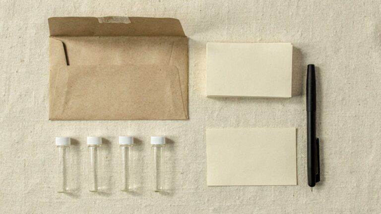 Overhead still life on cream linen: an open kraft padded envelope, four small empty glass sample tubes with white caps, blank cream cards and a black pen