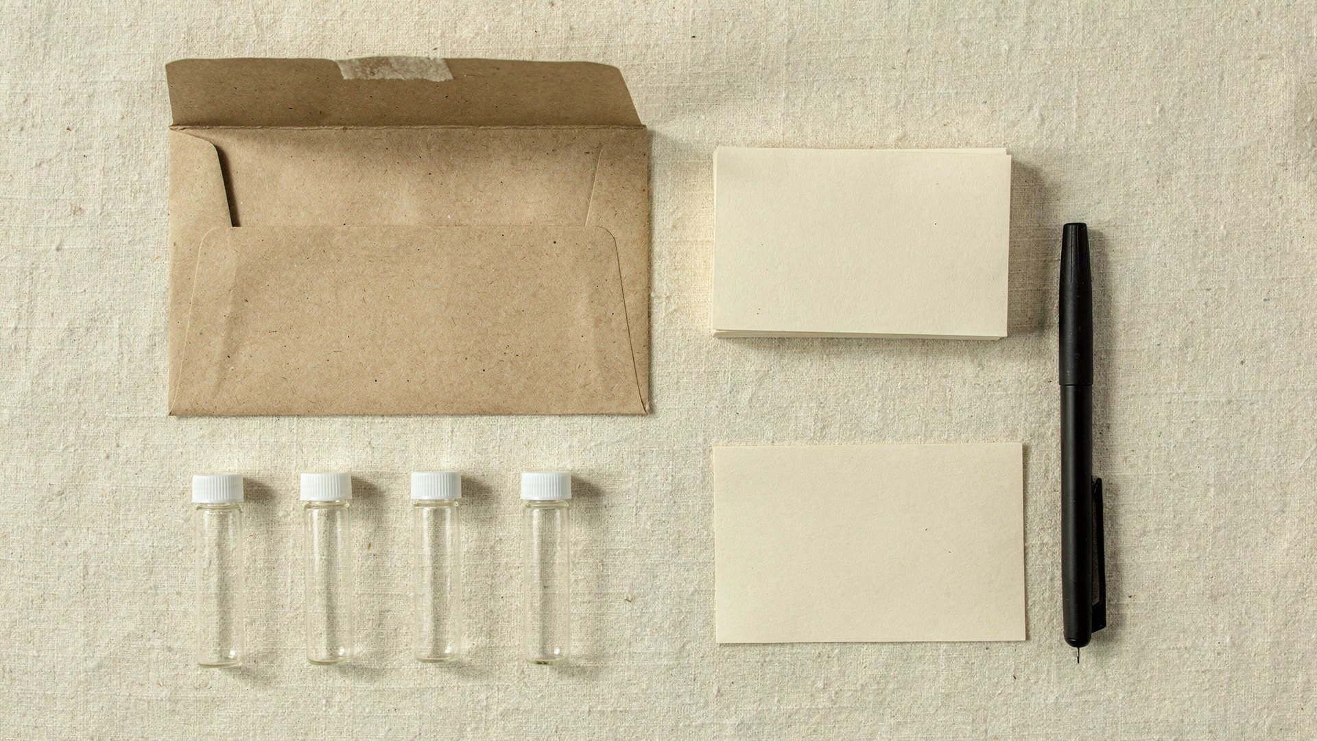 Overhead still life on cream linen: an open kraft padded envelope, four small empty glass sample tubes with white caps, blank cream cards and a black pen