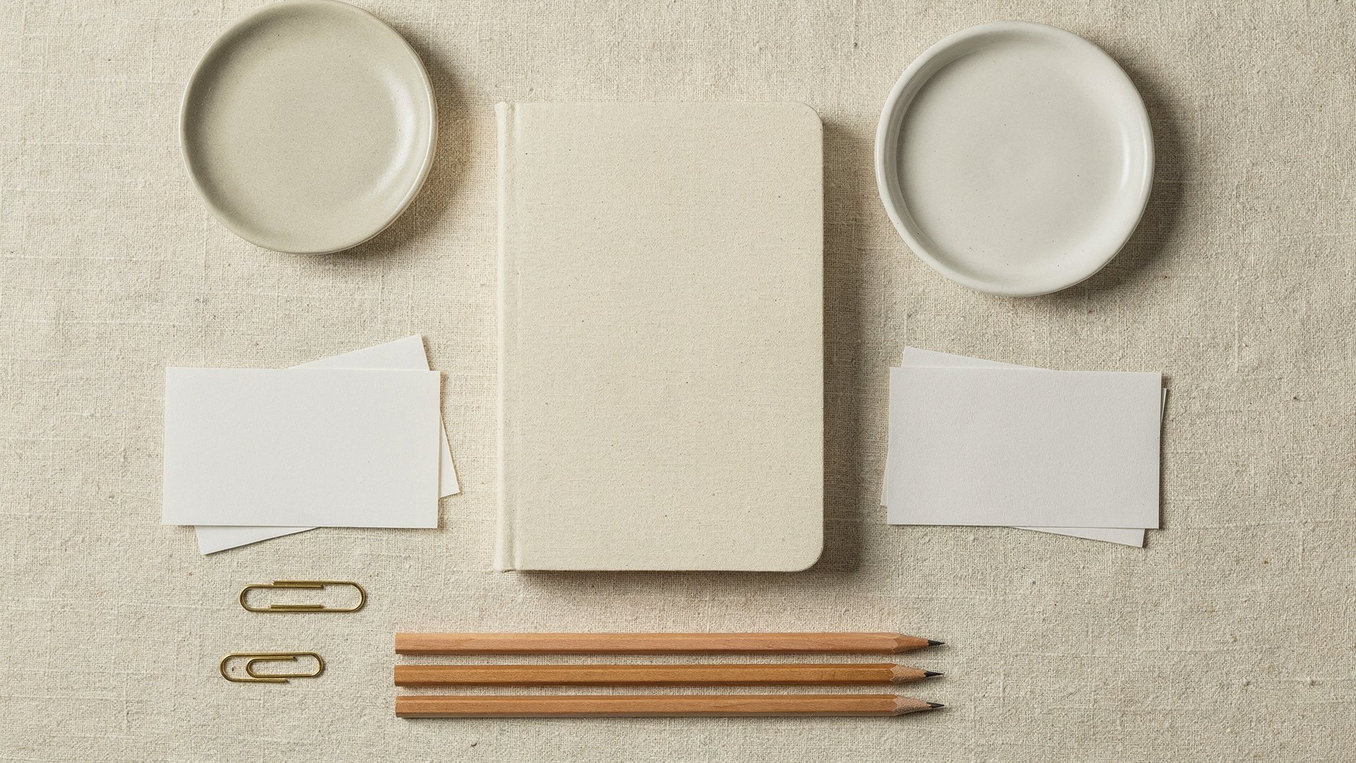 Overhead flat-lay on cream linen: a blank cream notebook, three wooden pencils in a row, blank white index cards, brass paperclips and two plain ceramic dishes