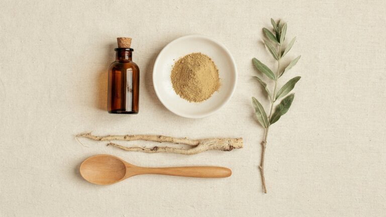 Overhead flat-lay on cream linen: an unlabelled amber glass bottle, a white dish of pale ochre powder, dried root pieces, a wooden spoon and a sprig of dried grey-green leaves