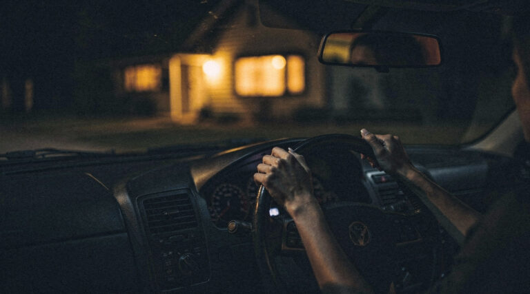 Hands resting on a steering wheel in a parked car at night with the lit house ahead, the pause before going in.