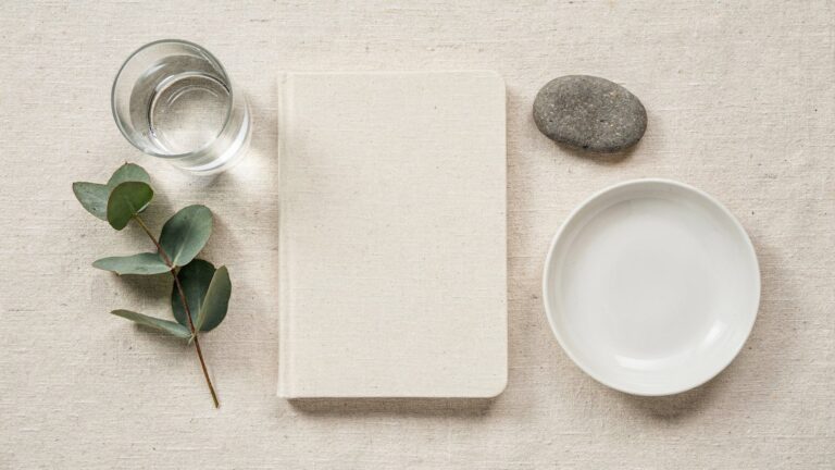 Overhead flat-lay on cream linen: a blank cream notebook, a glass of water, a smooth grey stone, a eucalyptus sprig and a plain white dish
