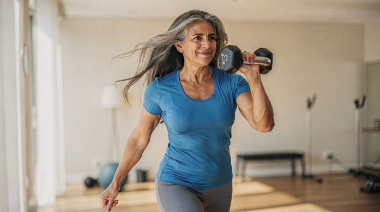 A woman with long grey hair lifts a dumbbell to her shoulder mid-stride in a bright home gym, smiling and strong