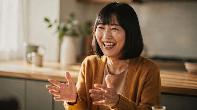 A woman with a black bob laughs with an open, delighted expression, hands mid-gesture, in a warm kitchen