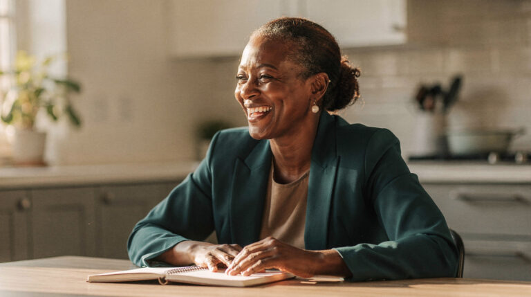 A woman in her late forties in a teal blazer laughs while sitting at a kitchen table with an open notebook, morning light behind her