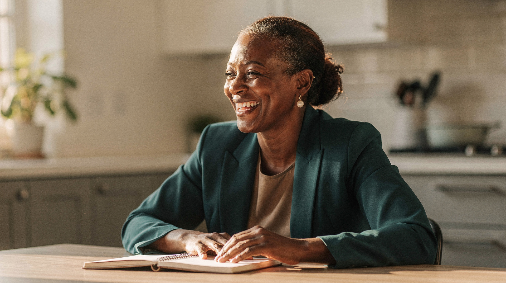 A woman in her late forties in a teal blazer laughs while sitting at a kitchen table with an open notebook, morning light behind her