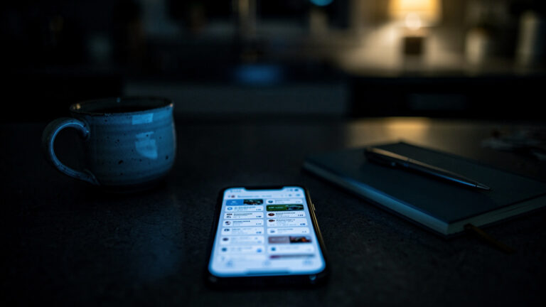 A phone glowing with open browser tabs on a dark kitchen counter at night beside a cold mug of tea and a closed notebook with a pen