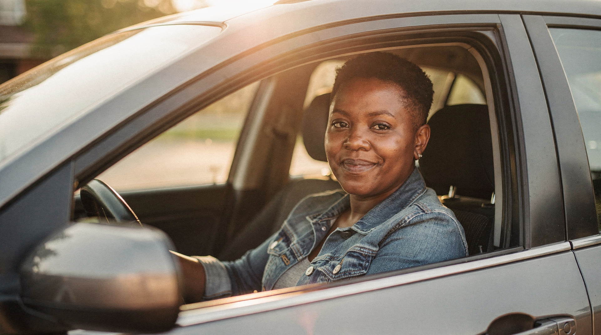 A woman with a short natural crop sits in a parked car with the window down, calm and smiling softly in golden light