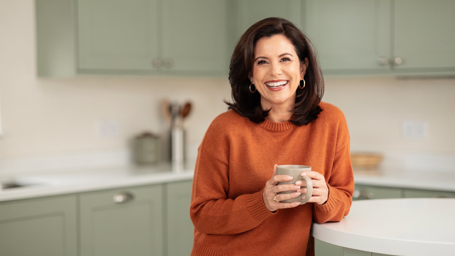 Brunette woman in her mid-forties laughing in her kitchen with a mug, relieved to know whether you can get pregnant during perimenopause