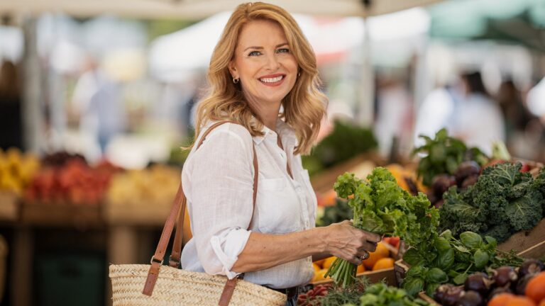Smiling strawberry-blonde woman in her late forties choosing fresh greens at a farmers market, thinking about foods that fight hot flashes
