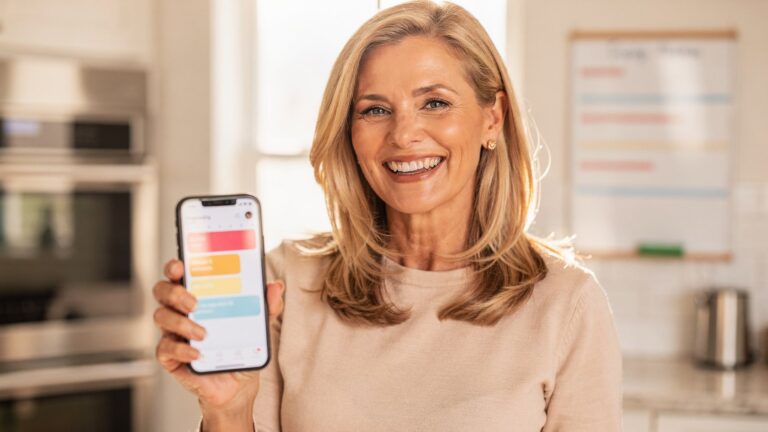 Smiling woman in her kitchen holding a phone displaying a color-coded shared family schedule app