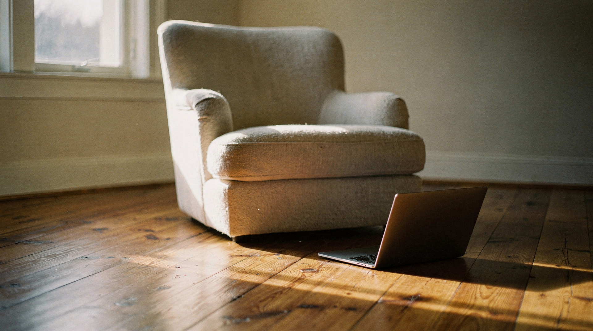 An empty armchair by a window with a closed laptop set down on the floorboards beside it in late afternoon light