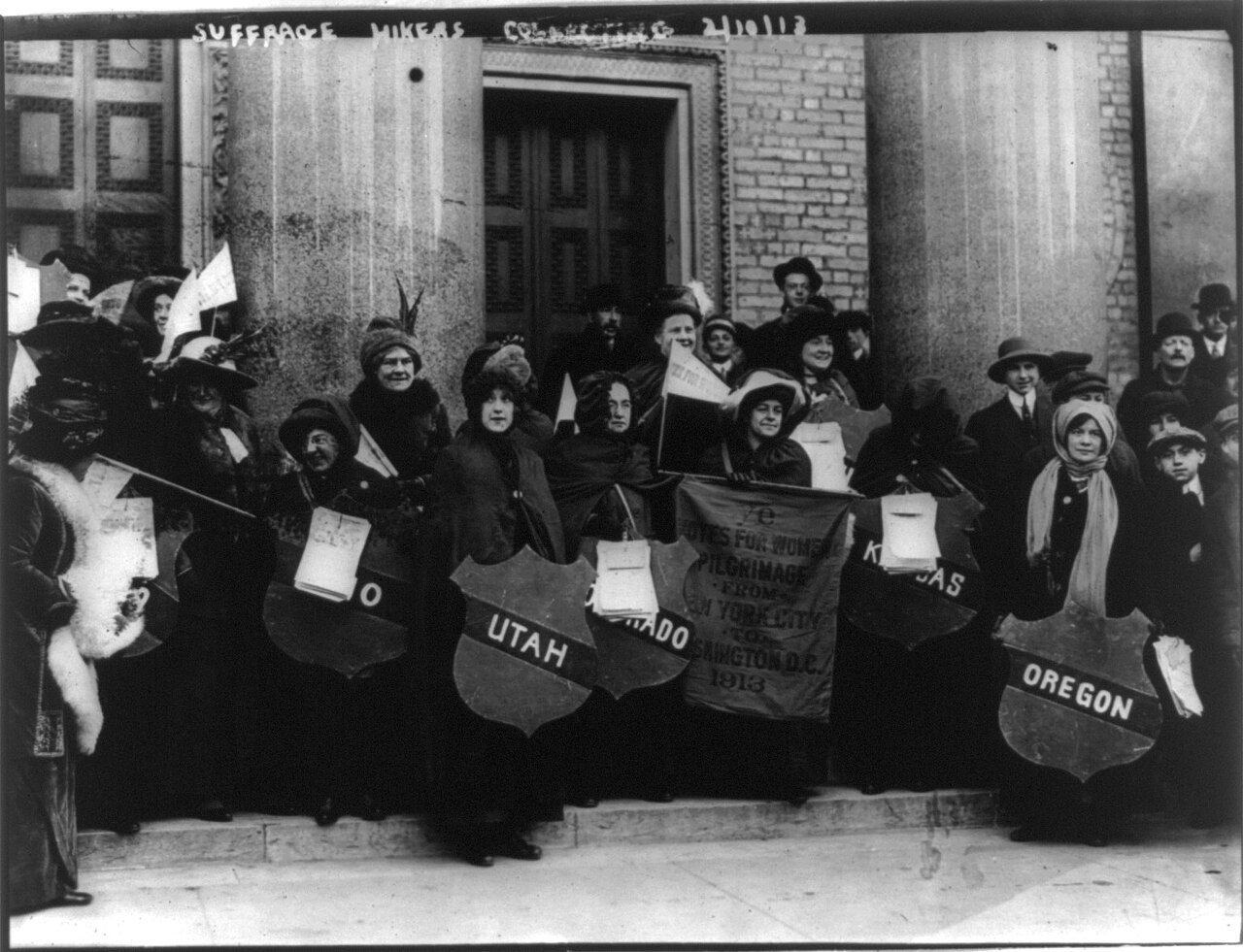 A group of suffrage hikers in winter coats holding state banners on the steps of a building, 1913, black and white
