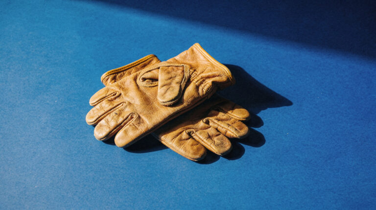 A worn pair of tan leather work gloves lying on a deep blue surface in hard sunlight, casting a long shadow