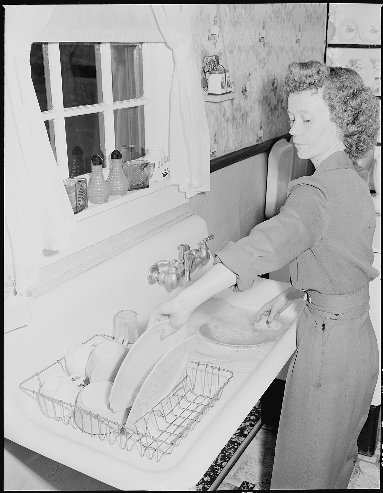 A young woman washing the breakfast dishes at the kitchen sink in a mid-century American home