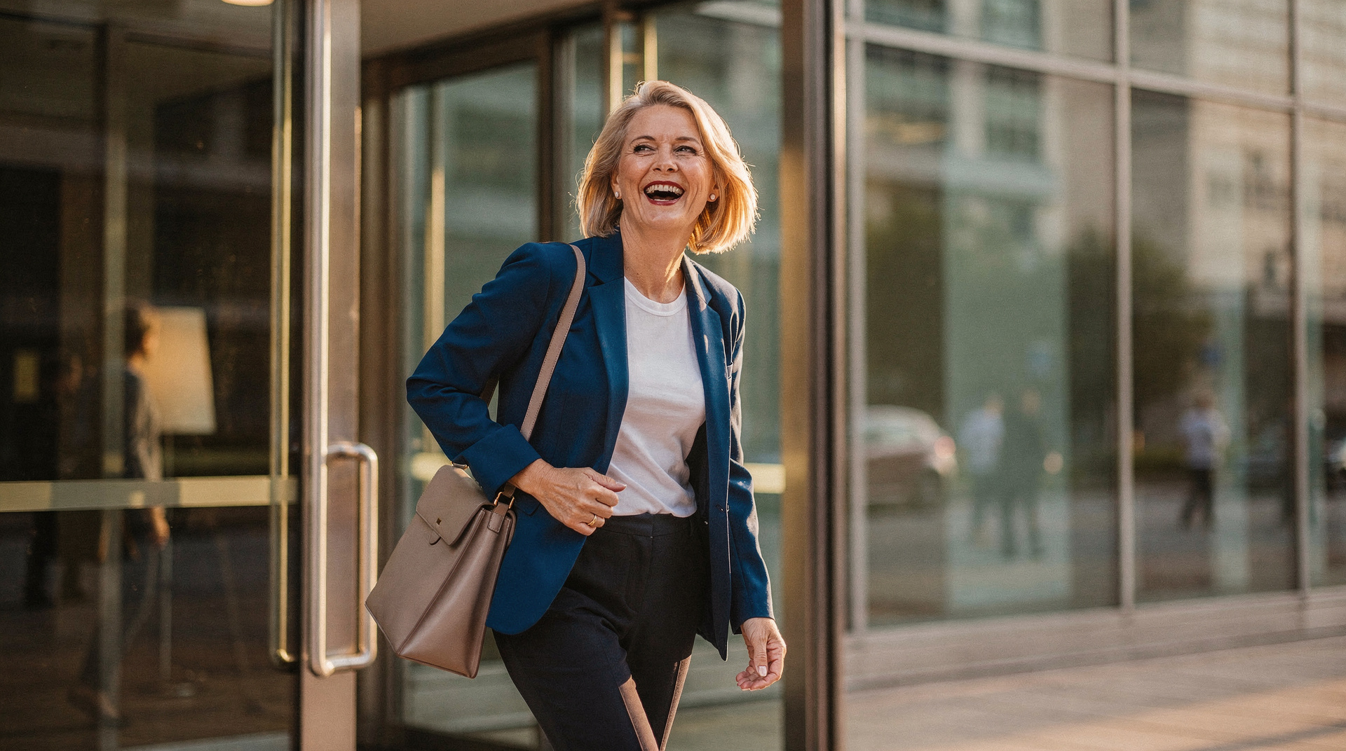A blonde woman in a cobalt blazer laughing as she walks out of an office building into late afternoon sunshine