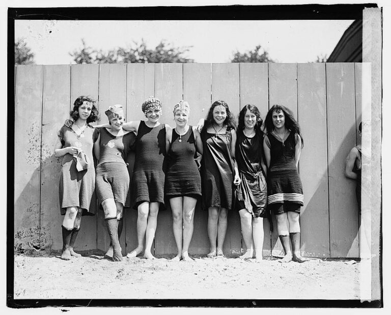 Who invented the weekend: seven young women in 1920s bathing suits standing arm in arm against a wooden fence, laughing, black and white