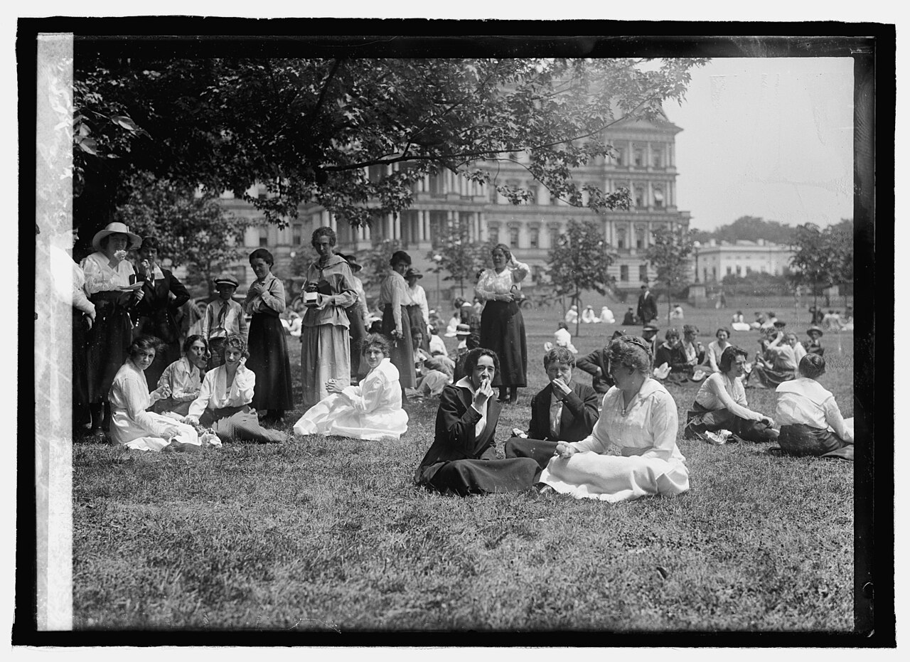 Women government clerks in long skirts and hats eating lunch together on a lawn in Washington, around 1918, black and white