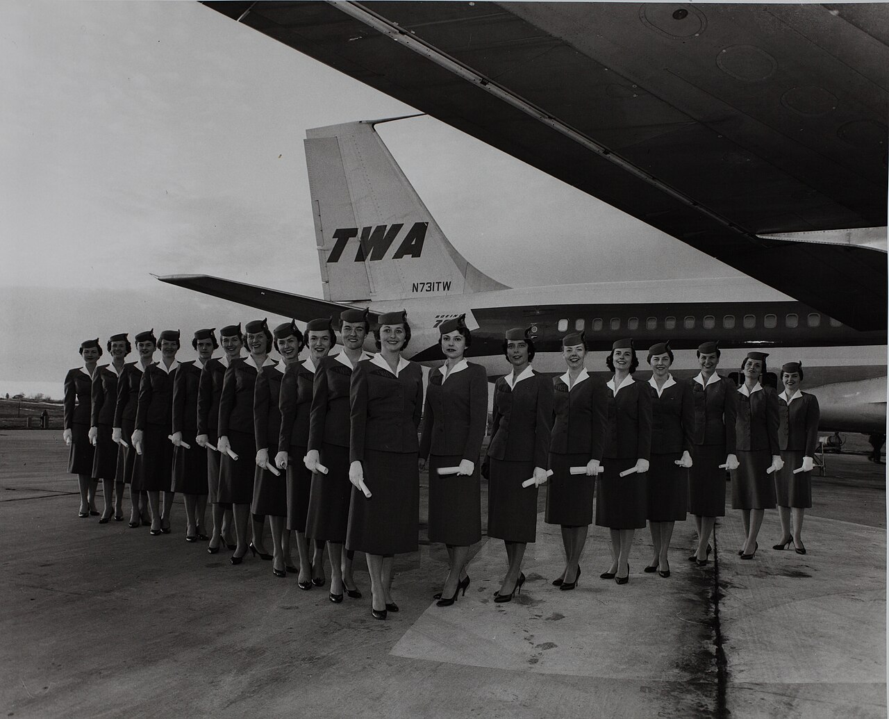 A long line of TWA stewardesses in identical uniforms standing in front of a jet, black and white