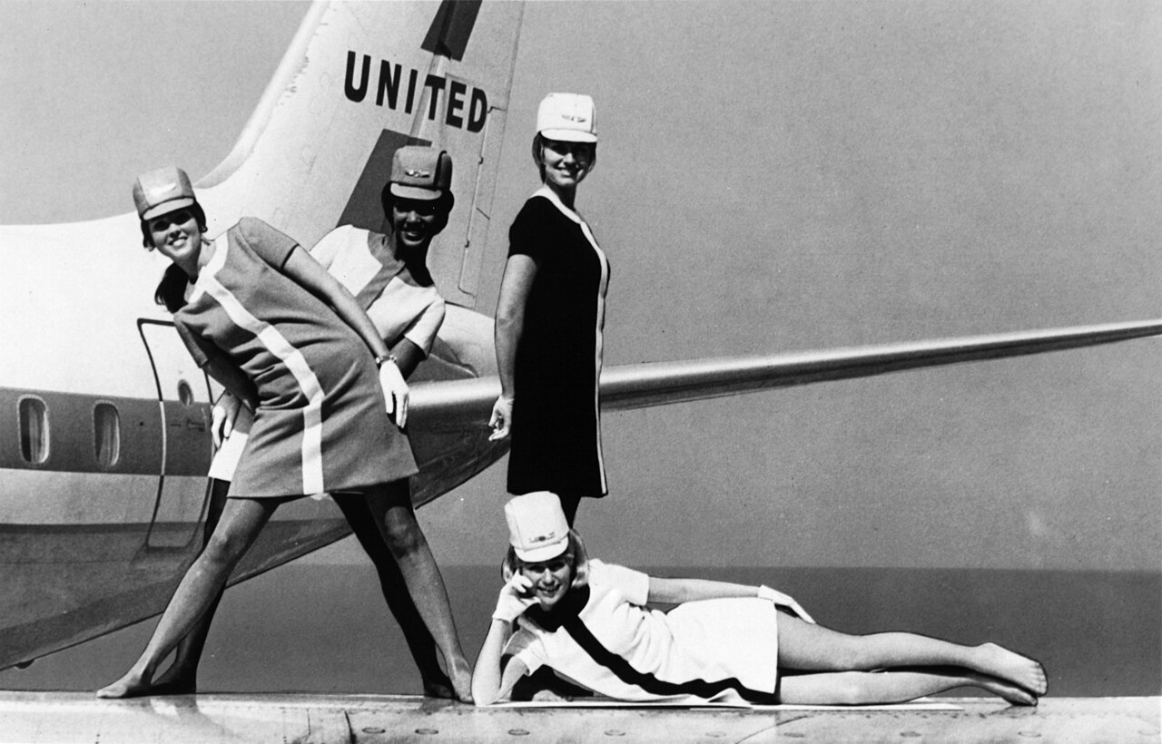 What is emotional labor: four United Airlines stewardesses posing and smiling in front of a jet tail, black and white, 1968